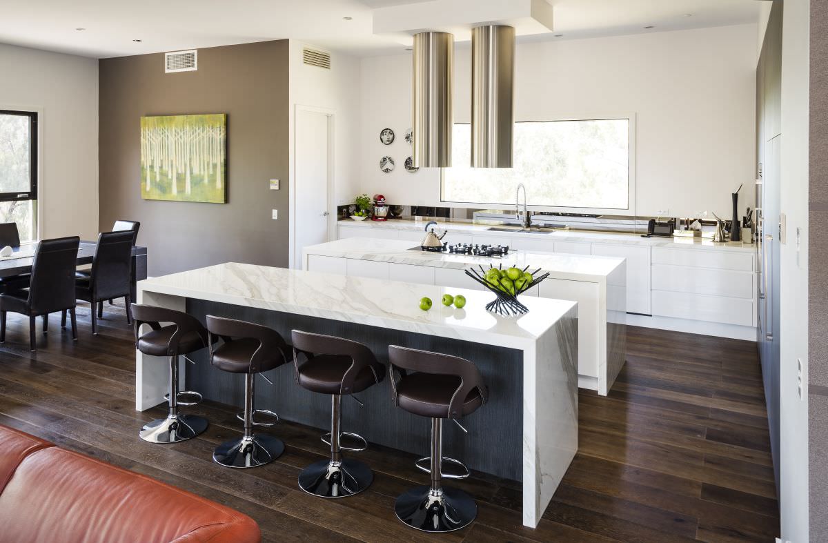 آشپزخانه مدرن و لوکسی که دارای کابینت جزیزه ای، بار، کابینت های سفید، هود استوانه ای و کفپوش چوبی می باشد